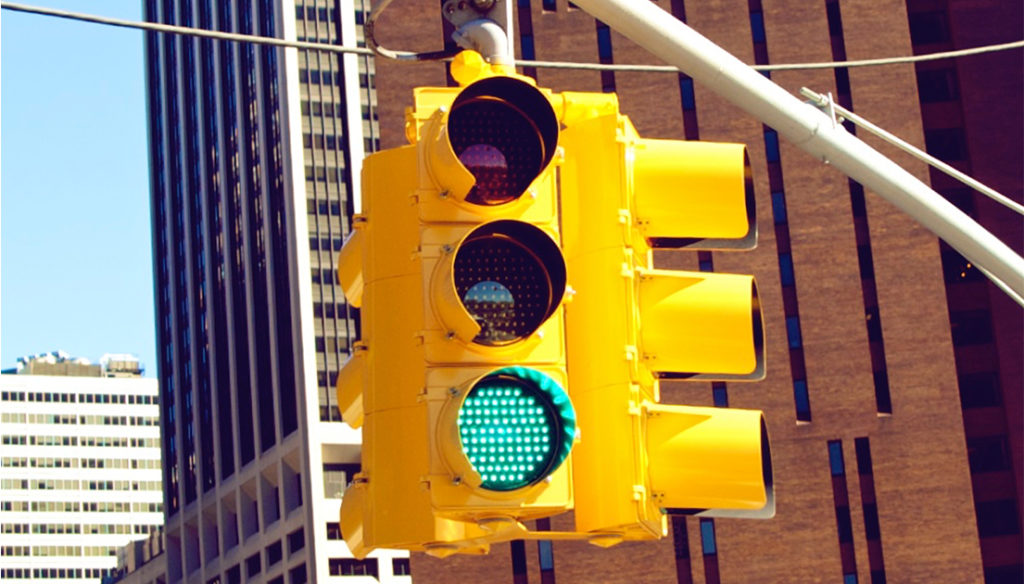 One side of a four-way traffic light flashes green to the traffic below. The traffic light hangs from a gray horizontal pole with telephone wires hanging overhead. The bases of two tall buildings are visible behind the traffic light. The first is made of brown brick and has long vertical windows. The second one is white with horizontal windows on the bottom and vertical ones near the top. The light blue sky and a smaller white building are visible behind the second building. The concept of the image is to use personality tests before giving candidates the green light to move forward in the hiring process.