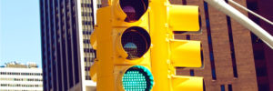 One side of a four-way traffic light flashes green to the traffic below. The traffic light hangs from a gray horizontal pole with telephone wires hanging overhead. The bases of two tall buildings are visible behind the traffic light. The first is made of brown brick and has long vertical windows. The second one is white with horizontal windows on the bottom and vertical ones near the top. The light blue sky and a smaller white building are visible behind the second building. The concept of the image is to use personality tests before giving candidates the green light to move forward in the hiring process.