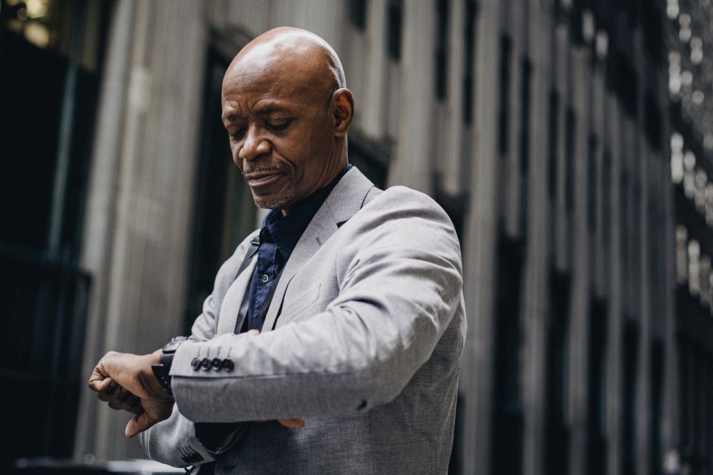 A bald black man with gray facial hair wears a light gray blazer with a navy blue collared shirt underneath. The city is an out-of-focus backdrop. The man furrows his brow as he checks his wristwatch, portraying patience or perhaps impatience. 