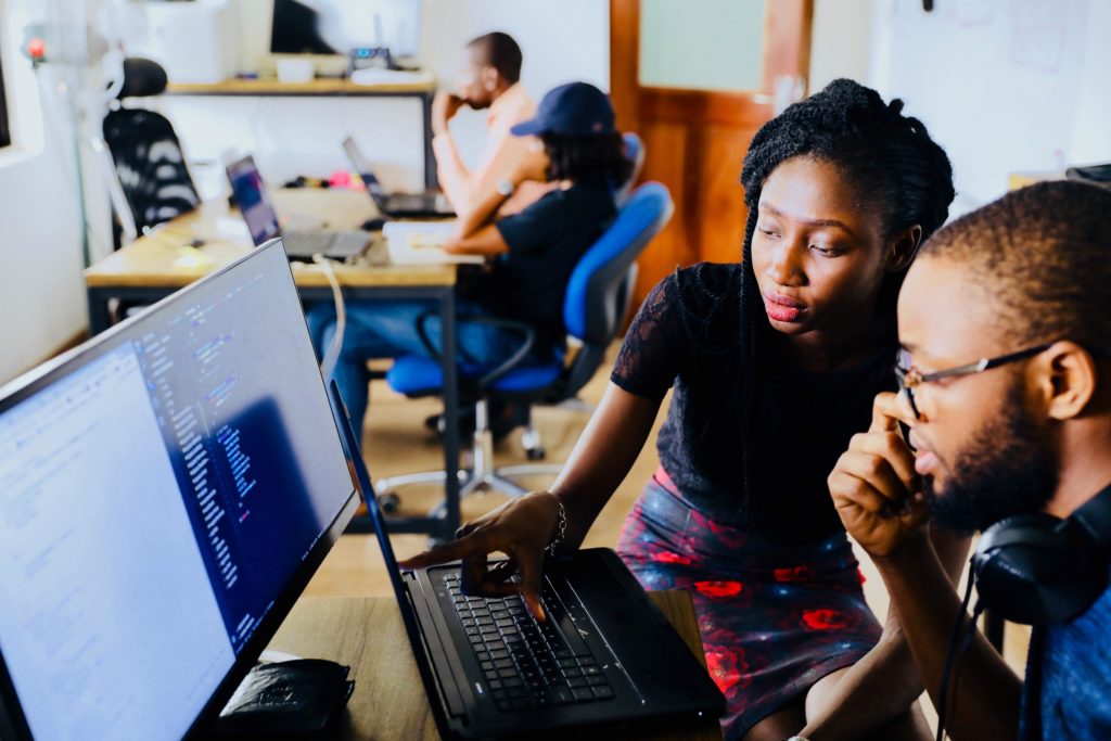 Two artificial intelligence professionals, a Black woman with locs and a Black man with a shaved head, beard, and glasses, are sitting in front of dual computer monitors, and the woman is gesturing as if she is giving instructions about coding. 