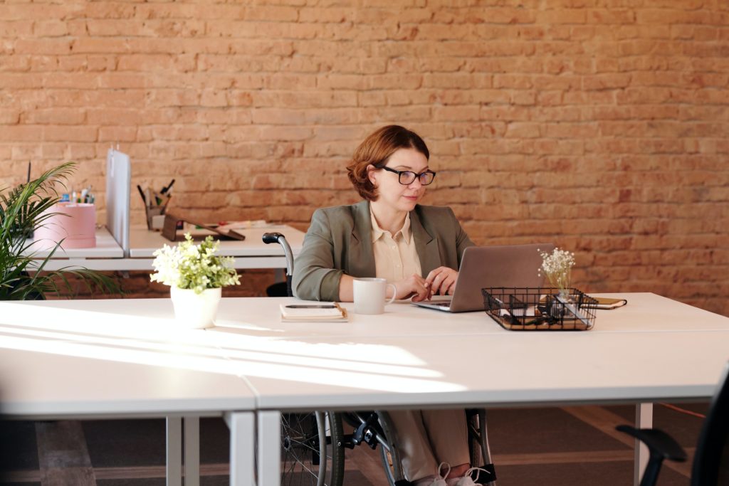 A woman with short red hair wearing a sage-colored blazer over an ecru blouse holds a remote coaching meeting via laptop. She wears square black eyeglasses and uses a wheelchair. She sits at a white table in front of a brick wall. On the tabletop is a white coffee mug, a notebook and pen, two small flower vases, and a basket of office supplies.