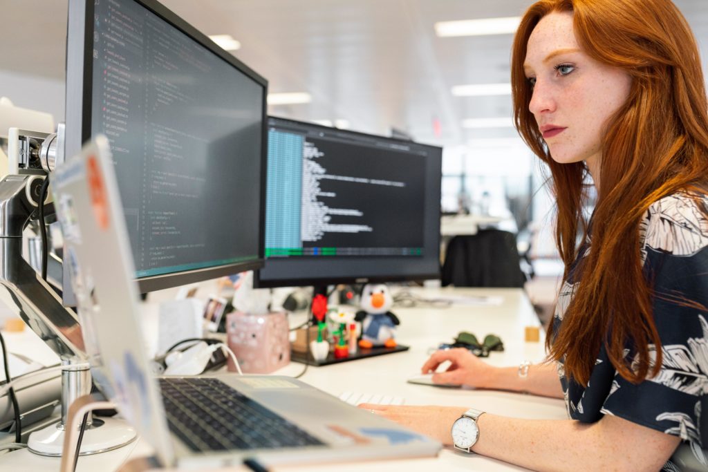 A female software developer, or artificial intelligence professional, with long red hair who is wearing a navy blouse with a white leaf print, a silver watch, and a silver bracelet sits at a desk in an open-plan office. She has two computer monitors and a laptop in front of her, along with some small tchotchkes and a box of tissues, and she is writing code for AI. 