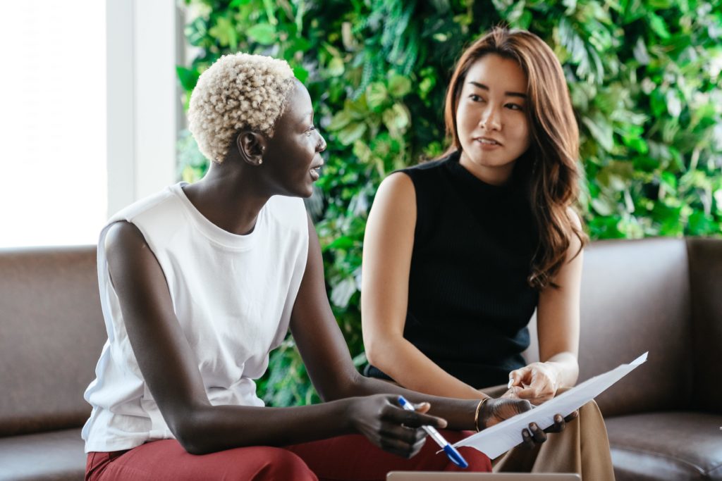 Two female artificial intelligence professionals, one Black with cropped platinum hair (left) and one Asian with long brown hair (right), sit on a sofa having a leadership development conversation. The woman on the left is holding a piece of paper and a pen. A tablet is on a table in front of them, and a wall of greenery is behind them.