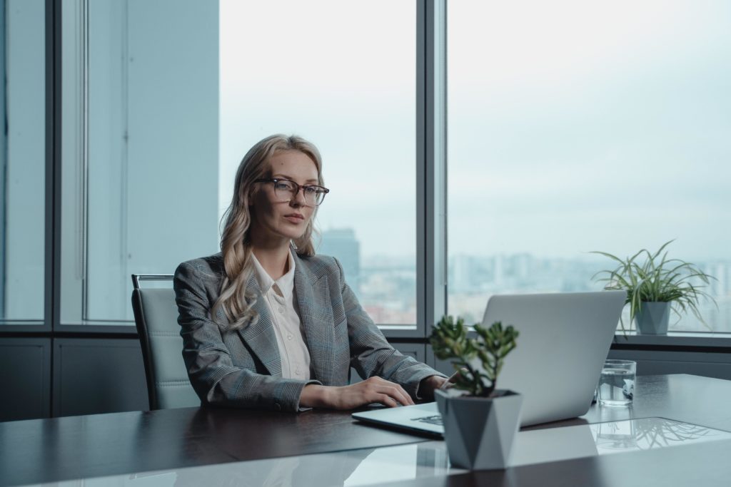 A blonde white woman CEO wearing glasses and a gray plaid blazer sits at a conference table working on a laptop. A large window with a foggy city skyline is behind her.