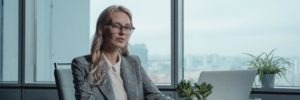 A blonde white woman CEO wearing glasses and a gray plaid blazer sits at a conference table working on a laptop. A large window with a foggy city skyline is behind her.