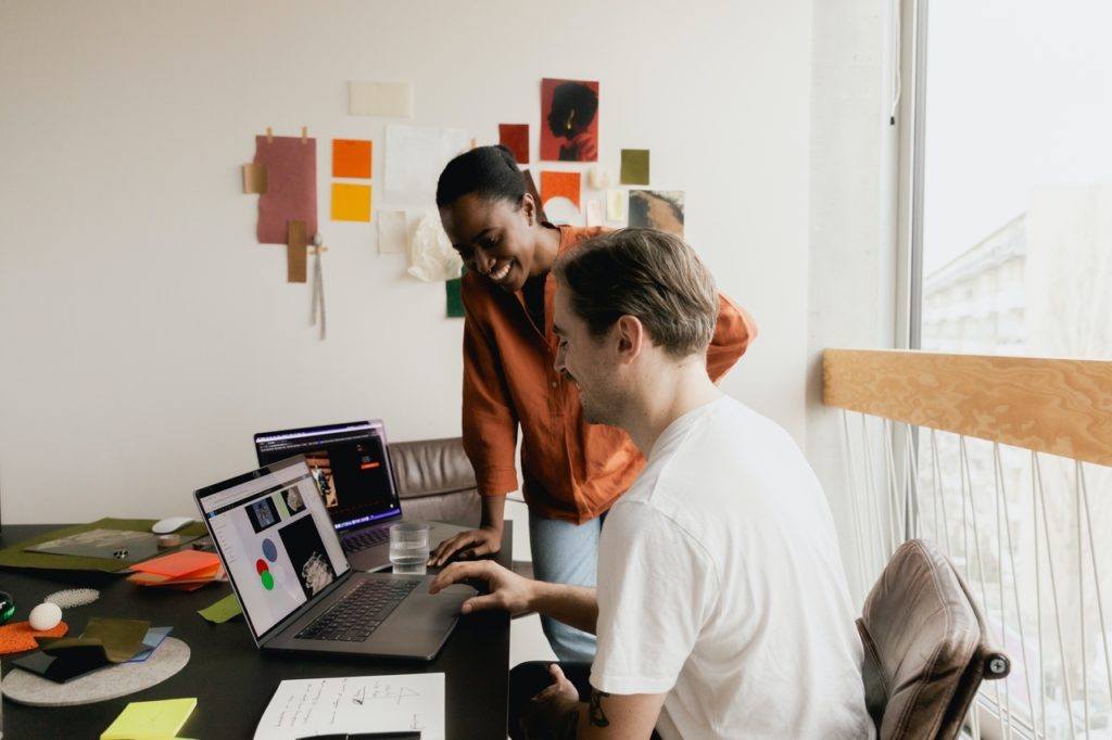 Illustrating the concept of employee engagement, two professionals smile as they collaborate on a project. One is a white man in a white T-shirt with a mustache. He is seated at a big black desk and is clicking on the touchpad of his laptop computer as the other, a Black woman in an orange blouse and blue jeans, leans against the table and watches him. They are in a brightly lit room with white walls and behind them is a large window facing the street.