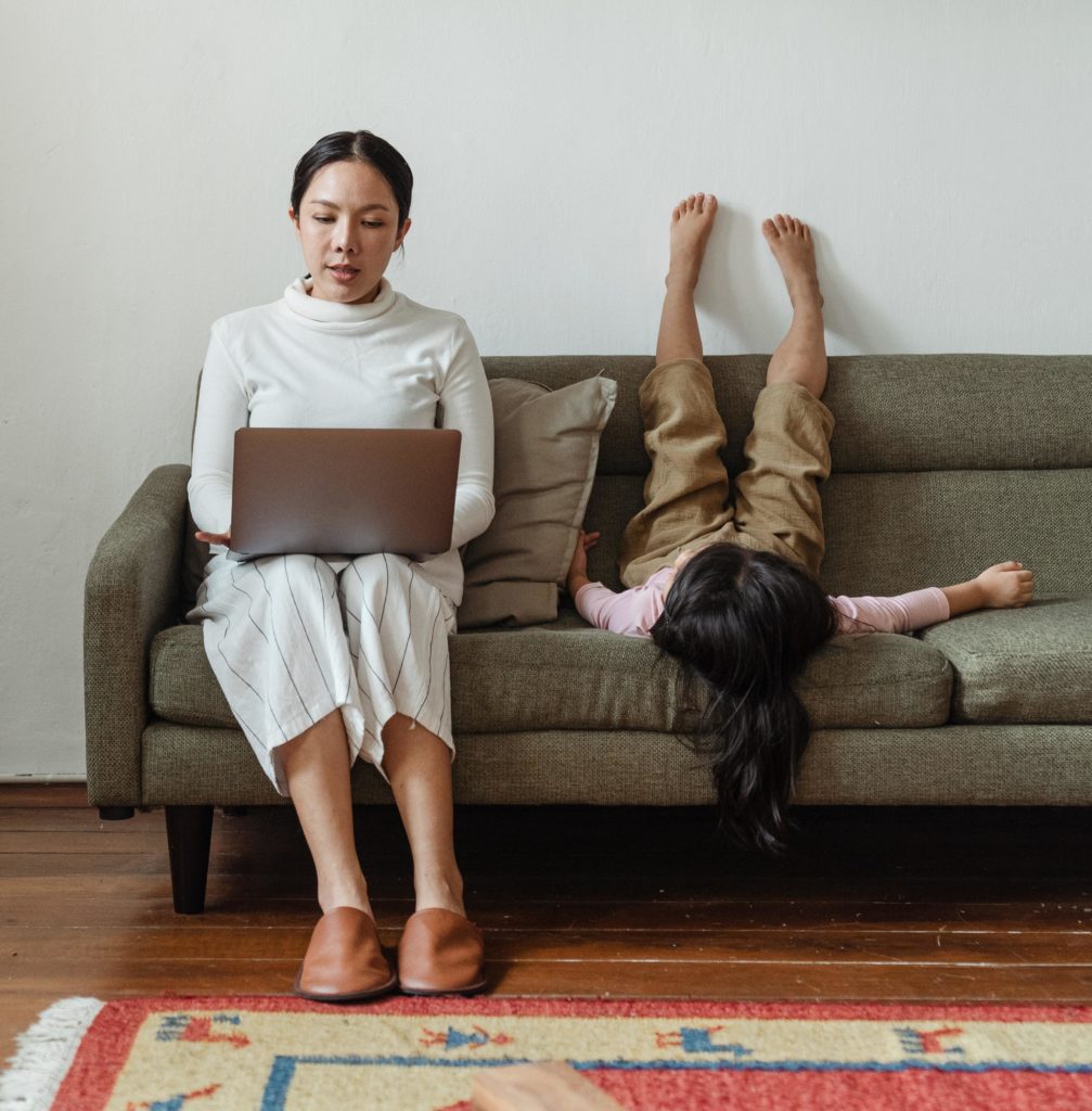 An Asian woman wearing a white turtleneck, pinstriped white gaucho pants, loafers, and a ponytail works on her laptop from a taupe-colored tweed couch. A white wall is behind her, and an orange, blue, and yellow rug lies on the floor in front of her. A long-haired child wearing a pink shirt and khaki shorts lies supine — legs stretched up the wall, looking restless — on the couch next to her.