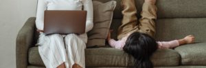 An Asian woman wearing a white turtleneck, pinstriped white gaucho pants, loafers, and a ponytail works on her laptop from a taupe-colored tweed couch. A white wall is behind her, and an orange, blue, and yellow rug lies on the floor in front of her. A long-haired child wearing a pink shirt and khaki shorts lies supine — legs stretched up the wall, looking restless — on the couch next to her.