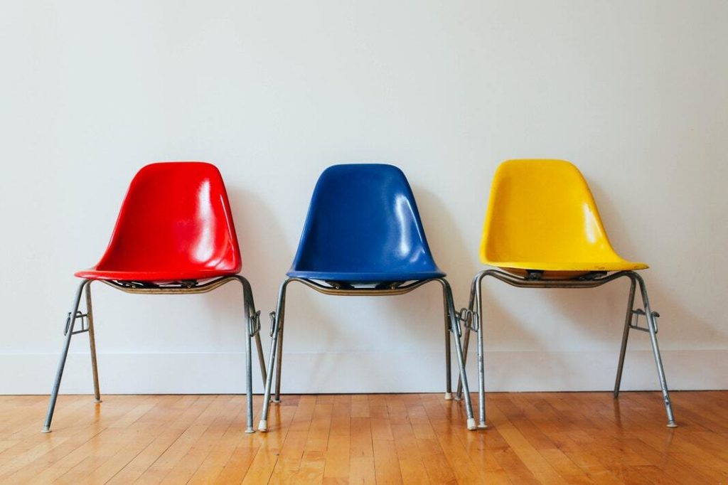 Signifying job candidate comparison, three molded plastic chairs with metal legs sit in a row against a white wall on a hardwood floor. The chair on the left is red, the chair in the middle is blue, and the chair on the right is yellow.