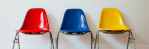 Signifying job candidate comparison, three molded plastic chairs with metal legs sit in a row against a white wall on a hardwood floor. The chair on the left is red, the chair in the middle is blue, and the chair on the right is yellow.