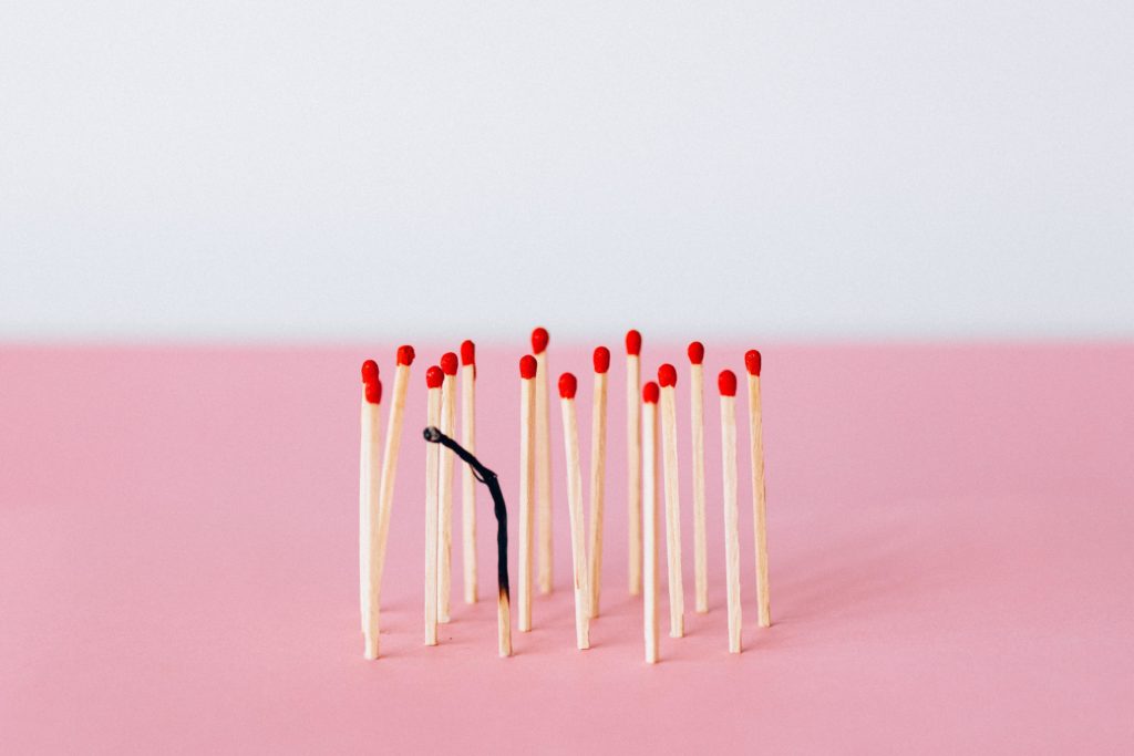 Numerous wooden matches with red tips stand upright on a pink surface in front of a white wall. Symbolizing employee burnout in the workplace, one match is almost completely charred and burned out. The wood is frayed on the sides, and the match is curled over at the top.  