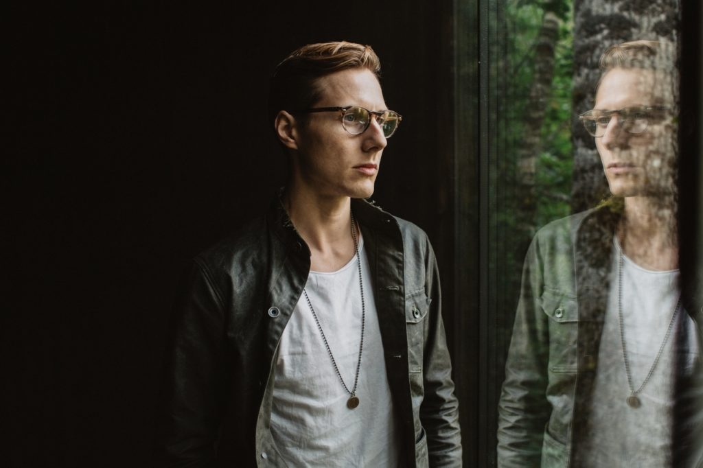 A solitary man stands in a pitch-black room and pensively looks out of a large window. He is wearing glasses, an unbuttoned jean jacket with a white tee shirt underneath, and a chain necklace with a penny on it. Symbolizing unconscious bias, the window is dominated by his reflection, which stares back at him.  