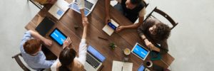 A group of employees and leaders sit around a conference table using laptops to discuss leadership development initiatives.