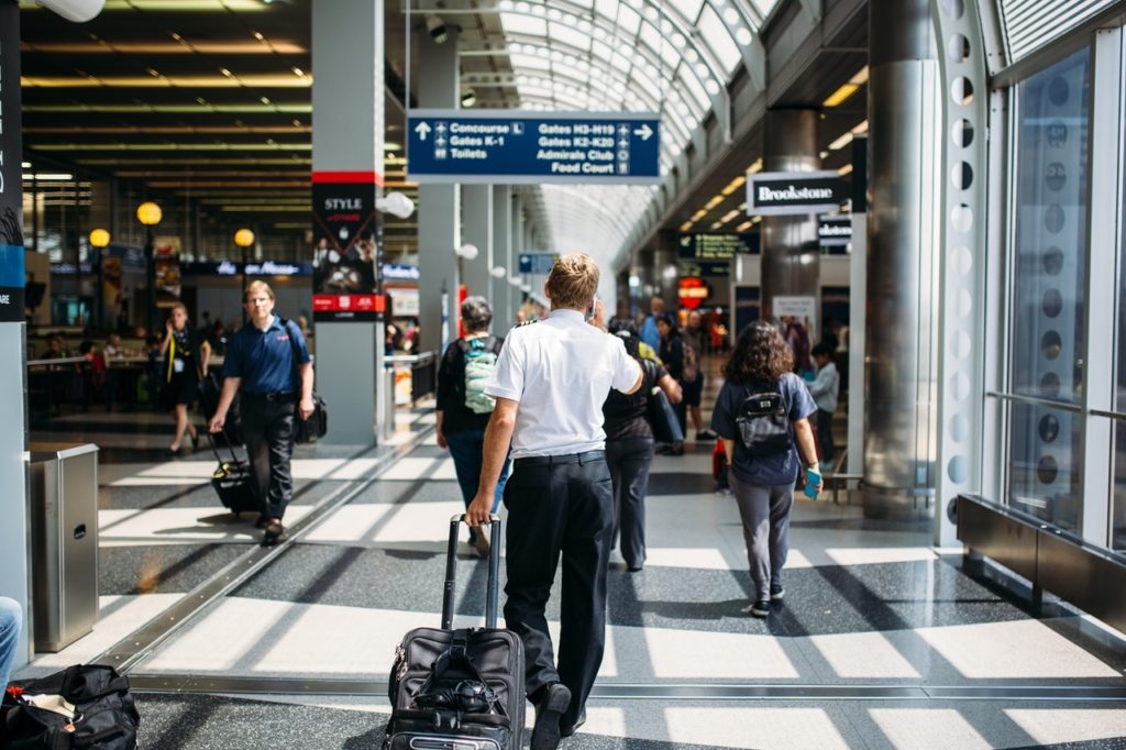 A pilot, an example of a high-stakes hire, walks through a busy hallway in Chicago O’Hare airport. His back is to us. He has short blond hair and is wearing a white pilot’s shirt tucked into black pants with black dress shoes. In his left hand, he pulls a rolling black suitcase. He holds a cell phone to his ear with his other hand. 