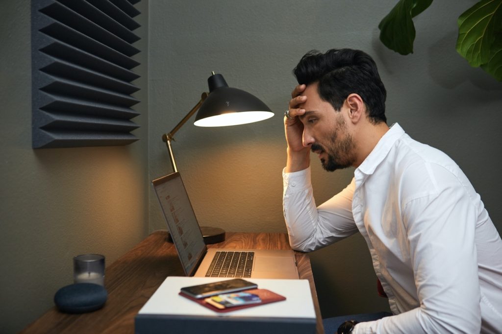 A dark-haired, bearded businessman wearing a white button-up shirt sits in front of a laptop computer illuminated by a black metal desk lamp. Experiencing interview fatigue, he rests his forehead in his hand. Also on his desk are a candle, a smartphone, and some books. Behind him is a houseplant. Accordian-shaped wall décor is mounted above his desk.