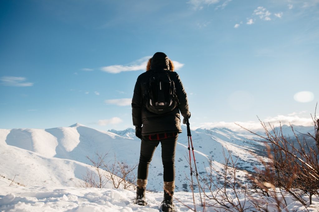 Signifying a leader embarking on a professional development journey with the guidance of a business coach who specializes in talent development, a mountaineer overlooks a snowy mountain range on a bright sunny day. Their back is to the camera. They are wearing a black jacket and backpack and holding two walking sticks with their right hand. 	