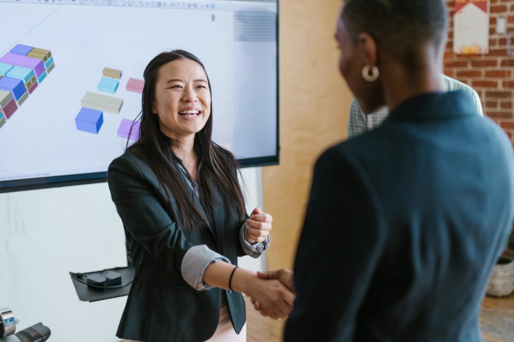 Two women wearing black blazers shake hands in front of a projector displaying business information. One of the women, who is Asian with long, straight hair, faces the camera and is smiling. The other woman, who is Black with a shaved head, has her back to the camera. 