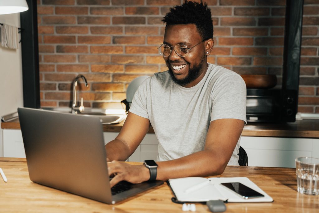 A Black remote worker with a beard and short hair smiles broadly while working from home using a laptop set up at a butcher-block kitchen counter. He wears round, wire-rimmed glasses, a gray T-shirt, an earring, and a smartwatch. In front of him lies a notebook, a smartphone, and a case holding wireless earbuds. A drinking glass sits left of his work area. An exposed brick wall is behind him.
