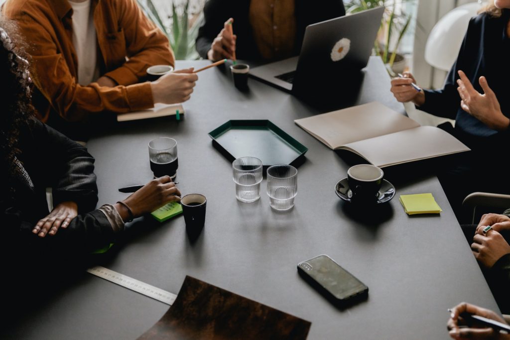 A racially diverse group of people sit around a conference table with notebooks, coffee, and laptops discussing how to weed out unqualified candidates. None of their faces are visible.