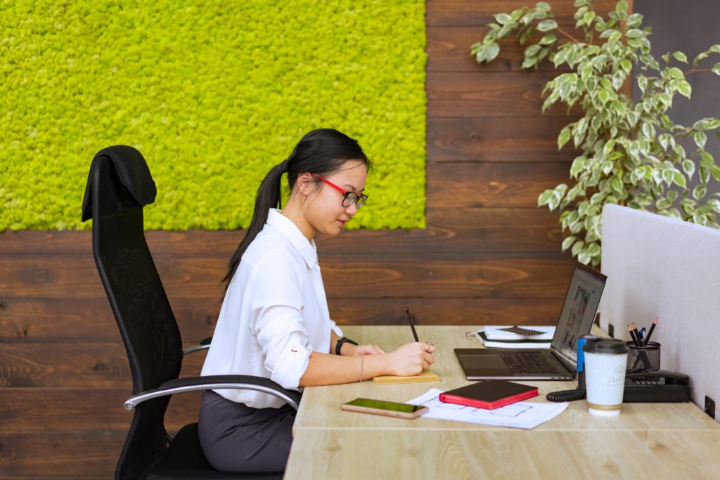 Why are so many people quitting their jobs? And what can organizations do to retain talent and keep employees engaged? A woman wearing a ponytail, red and black framed glasses, a white collared blouse, and gray slacks sits at a desk in front of a wooden wall adorned with foliage. She is working with a pen and paper, and a laptop, tablet, and smartphone sit on the desk surface in front of her. She also has a paper coffee cup and a corded phone.