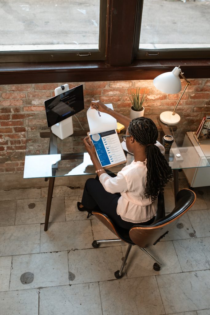 A hiring manager reviews a résumé on a clipboard, perhaps looking for résumé red flags. The hiring manager is a Black person with a ponytail and glasses who is wearing a white blouse, daisy bracelet, black slacks, and black high heels, and who is holding a pen in their right hand. The person is seated at a glass-topped desk with a computer monitor to the left and a cup of coffee to the right. A small cactus-like plant sits at the back of the desk, next to a white task lamp illuminating the area. The desk is positioned against an exposed brick wall.