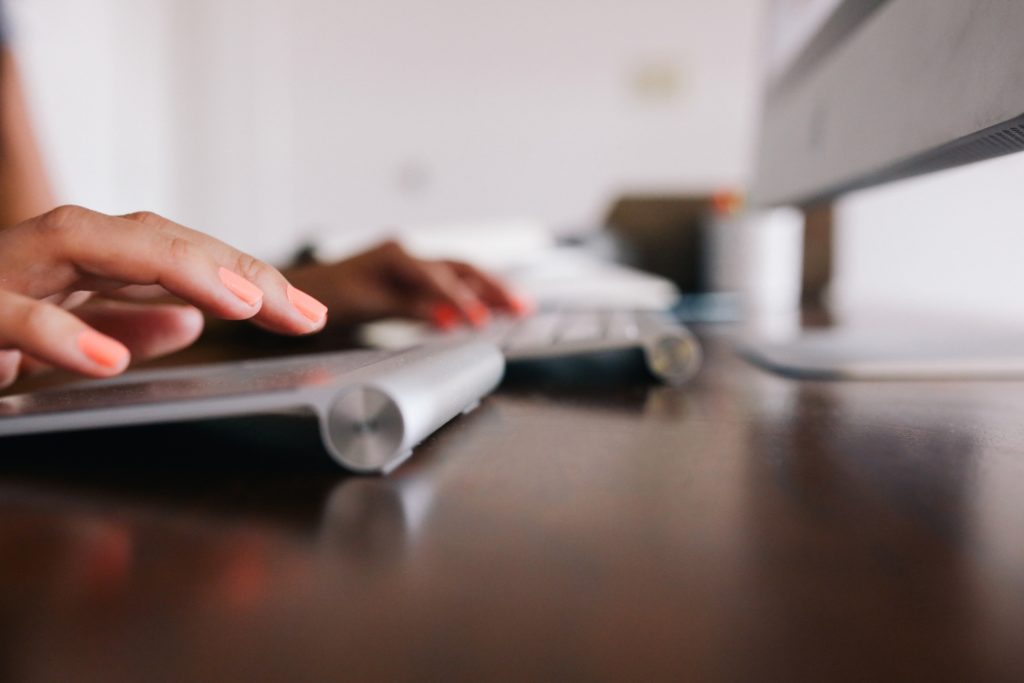 A close-up photo shows a person’s hands, which have coral-colored fingernails, as they use a keyboard and trackpad to write a rejection letter to a job candidate on a desktop computer.