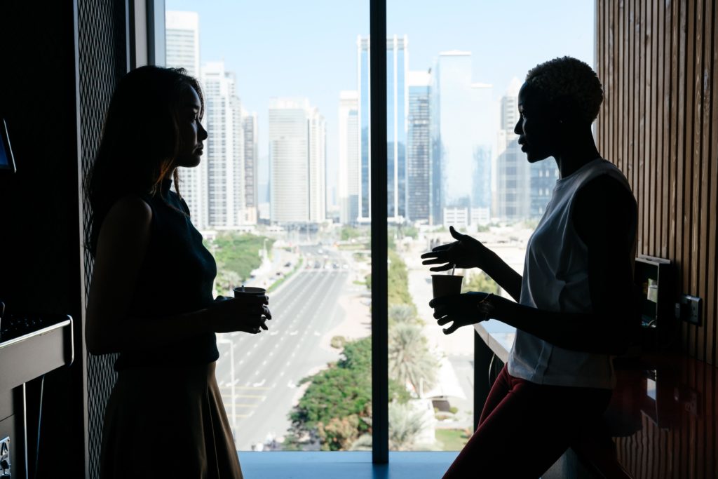 A manager and employee are standing in front of a window having a conversation. They are holding coffee cups and wearing business attire. Because they are backlit by the window, they are somewhat shadowy. The manager appears to be delivering feedback, signifying one of the challenges in becoming a manager. A city skyline and several lanes of highway traffic are visible through the window.