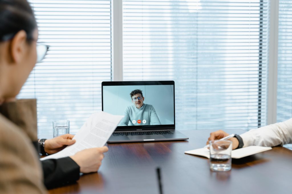 A laptop sits atop a conference table in front of a large window with open blinds. A virtual job interview is taking place. A job candidate with short, curly brown hair, a mustache, and square-framed black eyeglasses is visible on the laptop display. The interviewee is wearing a gray crewneck sweater and collared shirt and appears to be speaking. Three interviewers are visible around the conference table. One is looking at the laptop display, another is taking notes on the interviewee’s responses to the job interview questions, and the third is referencing a résumé or cover letter. Two glasses of water and a microphone are also visible on the table. The photo is intended to show principles for running a good job interview in action. 