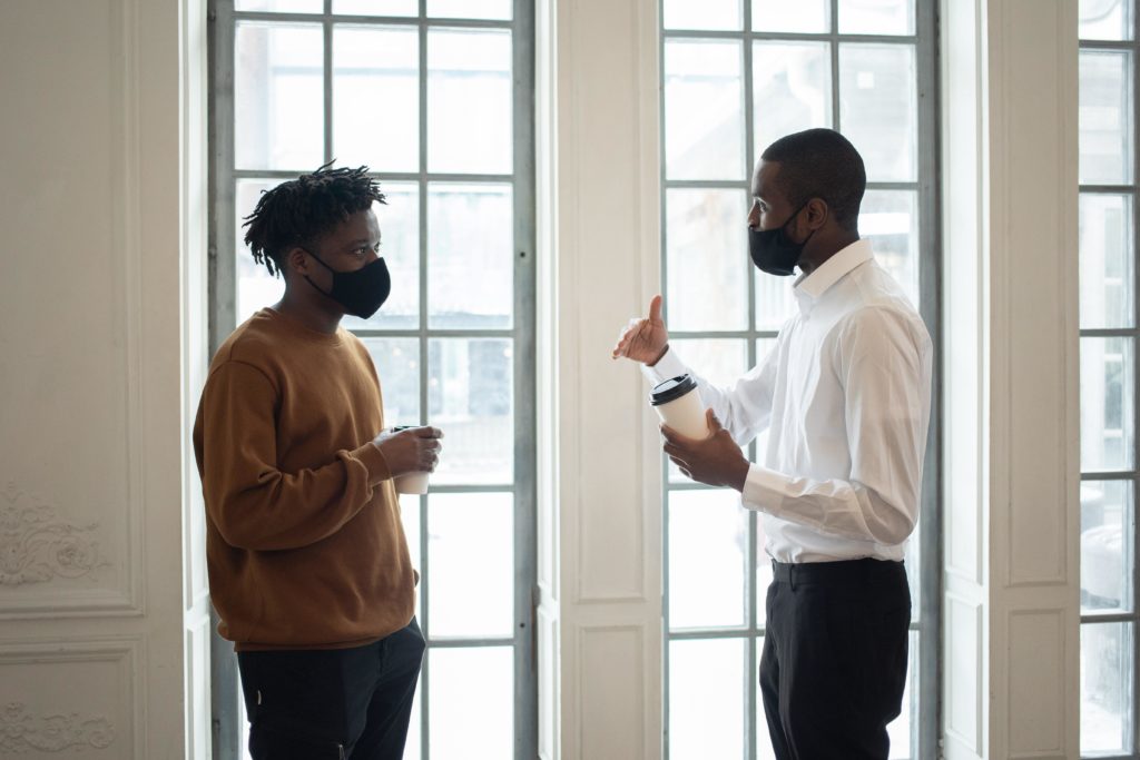 As part of the employee onboarding process, a manager and a new employee discuss the new hire’s development plan. They are standing in front of a large window, wearing business casual attire and face masks, and holding disposable coffee cups. 