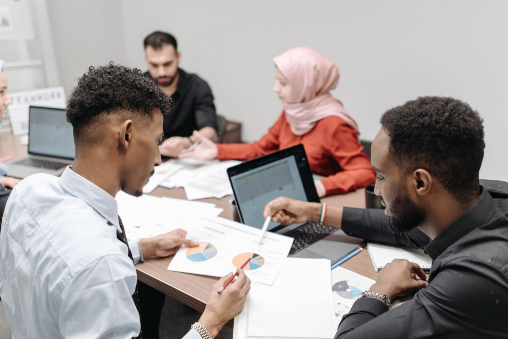 A group of five professionals seated around a conference table discuss business metrics, perhaps including the benefits of metrics-driven leadership development programs, such as enhanced team performance. A couple of laptops are atop the conference table, along with a mess of papers. The professionals are all wearing business attire. Two of the professionals in the foreground are gesturing at a piece of paper displaying pie charts.