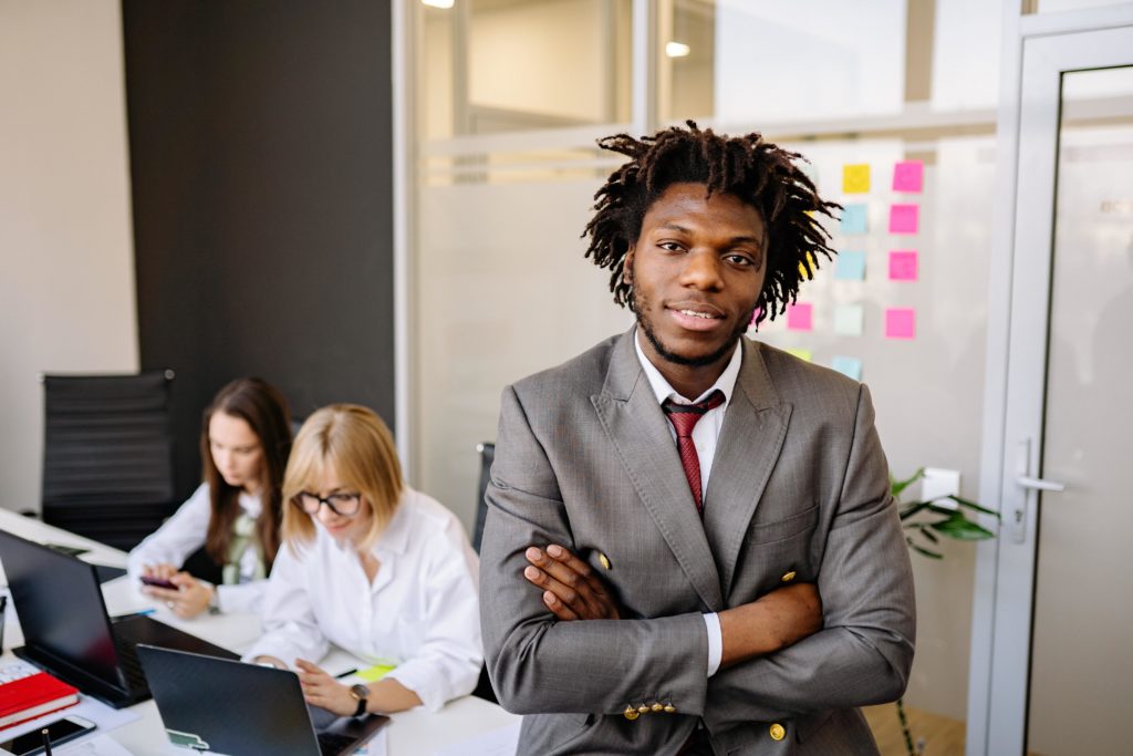 A person wearing a gray suit with a red tie and white shirt leans against a table with arms crossed, smiling slightly at the camera. Behind the person is a wall with a grid of colored sticky notes. Two other people work on laptops at the table. The people are participating in a leadership development program using 360-degree assessments and personality assessments for leader development.