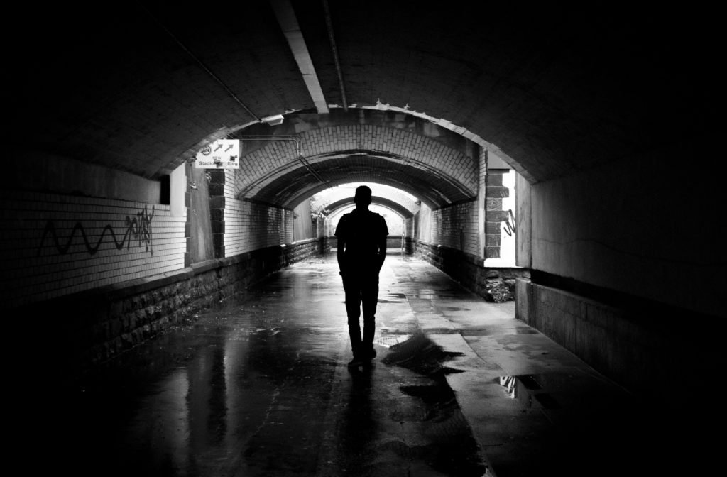 A silhouette of a man standing in a dark, damp, graffitied tunnel. The photo is in black and white, and its dark, gritty mood evokes the theme of the psychology of criminal behavior. The man’s anonymity represents the fact that, historically, little has been known about the role of personality in organized crime.