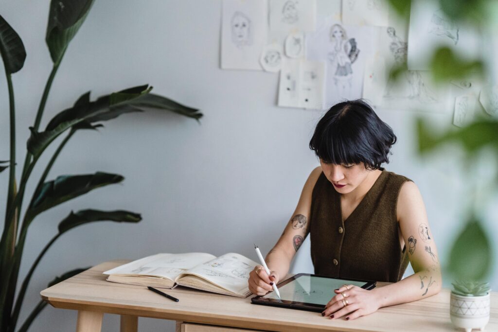 An employee with creative talent draws on a tablet at a pale wooden desk. She wears an olive-green sweater vest with brown buttons. She has short dark hair, tattooed arms, numerous rings on her hands, and dark nail polish. Next to her tablet is an open notebook with drawings in it. A pen lies on the desktop. Multiple drawings are posted on the wall behind her, and the desk is flanked by large houseplants. 