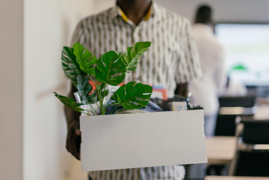 Employee turnover is on the rise with the Great Resignation, and many employers find they need to improve employee retention. This photo shows a person who has just quit his job. He is holding a box containing a plant and other assorted objects that might be found at an office desk. The former employee is wearing a patterned short-sleeved button-down shirt and is otherwise out of focus in the photo. 