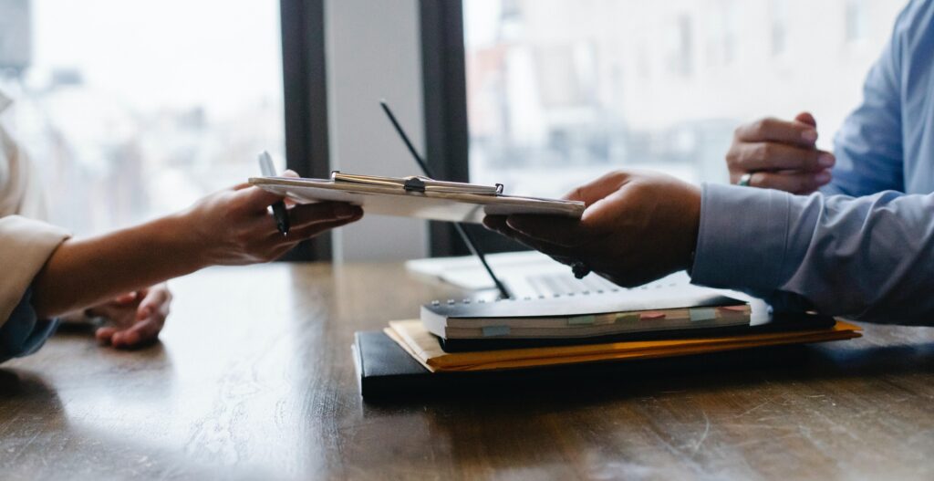 Two people, whose faces are not visible in the frame, exchange a clipboard over a wooden conference table. The people are preparing for structured job interviews. The conference table also holds a couple of notebooks, a binder, and some envelopes. There are large windows visible on the side of the table opposite the camera. 