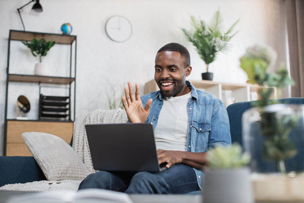 A remote worker dressed in casual attire is seated on a couch using a laptop. Facing the camera, the person is smiling and waving at the laptop’s built-in camera. The remote employee appears to be participating in virtual team-building activities, virtual team-building games, or virtual team development. The brightly lit room is out of the camera’s focus but contains several plants, a bookshelf, a wall-mounted clock, and a task lamp.