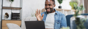 A remote worker dressed in casual attire is seated on a couch using a laptop. Facing the camera, the person is smiling and waving at the laptop’s built-in camera. The remote employee appears to be participating in virtual team-building activities, virtual team-building games, or virtual team development. The brightly lit room is out of the camera’s focus but contains several plants, a bookshelf, a wall-mounted clock, and a task lamp.