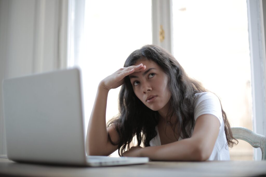 A medium-skinned person with long dark hair wearing a white T-shirt is seated at her desk. Her laptop is open in front of her, and a window is behind her. Her posture implies that she might be quiet quitting: her expression is frustrated, she is holding her hand over her forehead, and she is looking upward and to her left as if rolling her eyes.
