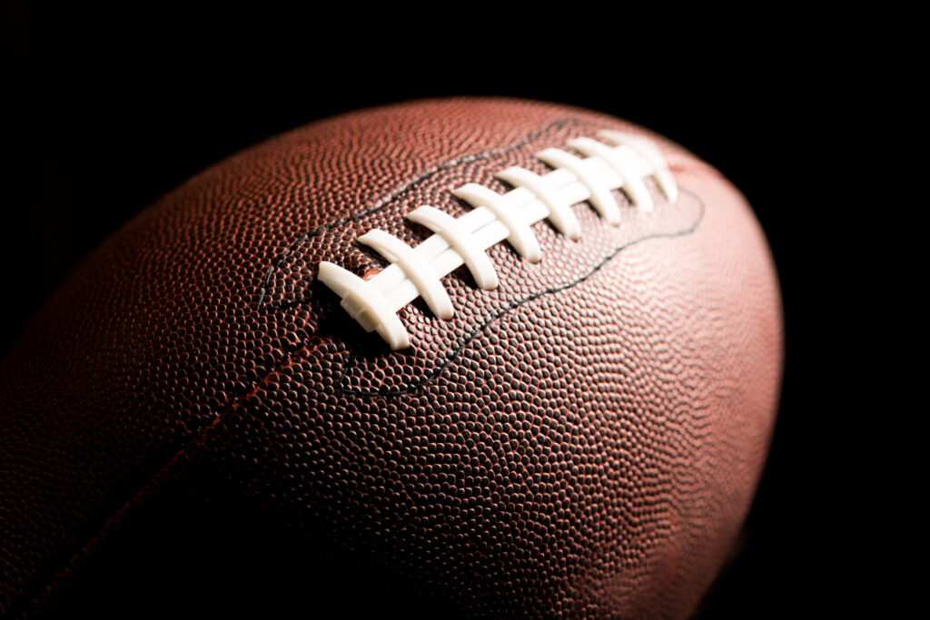 A close-up photograph of an American football against a black backdrop. Light is reflected on the stitched side of the ball. The image is used for a blog post about the reason why there are so few Black head coaches in the National Football League (NFL): bias in job interviews.
