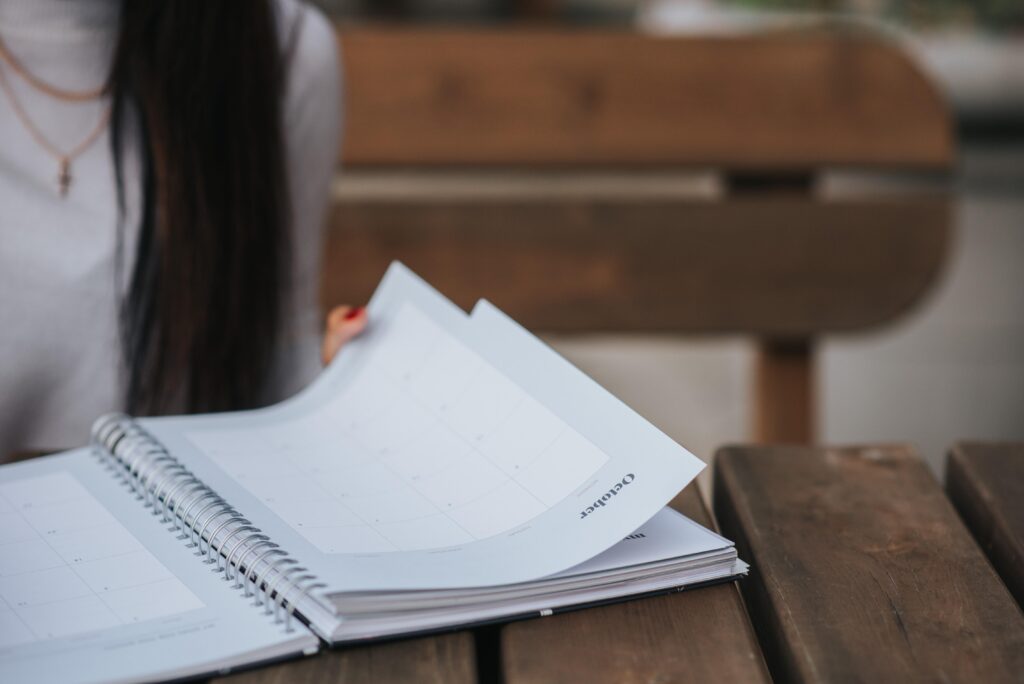 In this close-up photograph, a person with long dark hair and a gray sweater, whose face is not visible, is seated at a table. The person is leafing through a monthly planner on the table in front of them. The planner is open to the month of October. The implication for this blog is that the person might work in human resources and be planning a company holiday on Indigenous Peoples’ Day. 