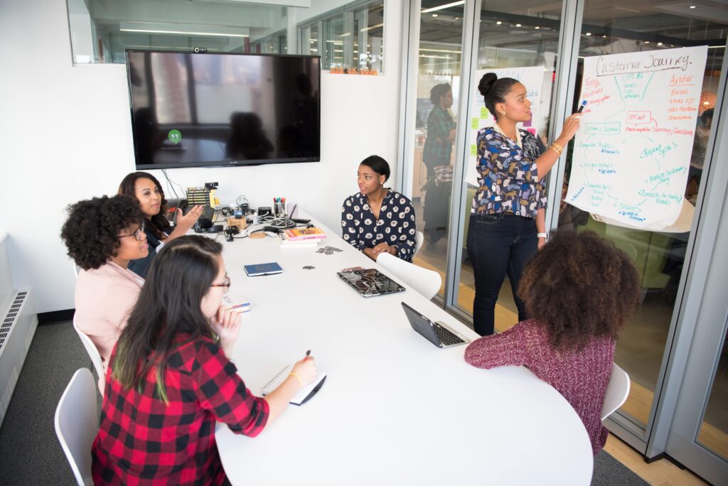 A group of multiethnic women are seated in white plastic chairs around a long white conference table with rounded edges. One stands in front of a poster attached to a floor-to-ceiling window. The poster displays notes about the customer journey, and she is pointing a marker at it. At the head of the table is a large monitor topped with a webcam and set to its screensaver. The table is topped with stacks of books, laptops, and various office supplies. Glass windows along two sides of the white-walled room reveal a larger office. Demonstrating team psychological safety, the women in the conference room appear to be engaged and working together to generate ideas.