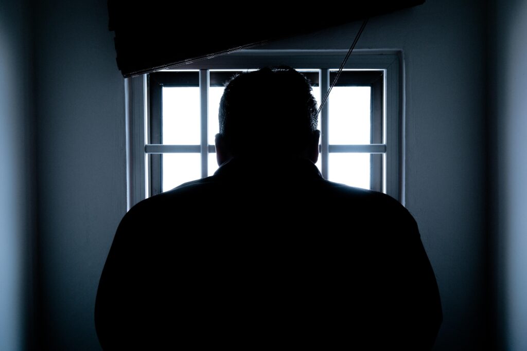 A person looks out of a barred window in a dark room or prison cell. Light is filtered through the window so only the person’s silhouette is visible, and the room appears shadowy. The image suggests that the personality of serial killers is ambiguous. 