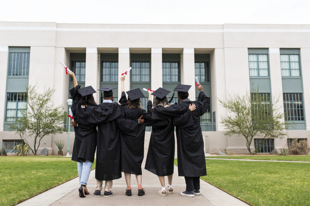 Five college graduates wearing academic gowns and mortar boards stand together facing an educational building while holding up their diplomas as if in salute. The photo illustrates a blog about the impact of personality, curiosity, intelligence, and conscientiousness on educational outcomes and career development. 