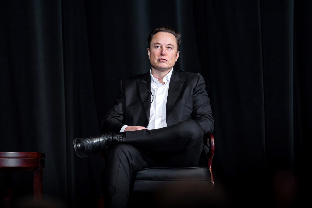 USAFA Hosts Elon Musk at an event in Colorado. Musk is dressed in a black suit, black boots, and is seated in a black chair with brown, wooden arm rests against a black backdrop.