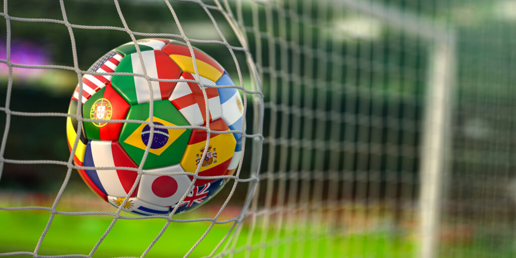 A soccer ball or football printed with various national flags bounces off of a goal net in a soccer/football stadium. The image illustrates a blog about the upcoming FIFA World Cup soccer/football games and how personality assessments can help predict the performance of professional athletes.