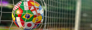 A soccer ball or football printed with various national flags bounces off of a goal net in a soccer/football stadium. The image illustrates a blog about the upcoming FIFA World Cup soccer/football games and how personality assessments can help predict the performance of professional athletes.