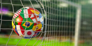 A soccer ball or football printed with various national flags bounces off of a goal net in a soccer/football stadium. The image illustrates a blog about the upcoming FIFA World Cup soccer/football games and how personality assessments can help predict the performance of professional athletes.