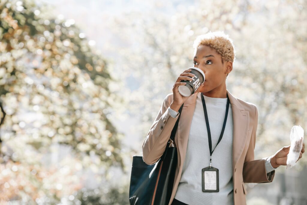 A person sips coffee from a takeout cup. In their other hand, they hold a takeout bakery item in a plastic bag. They are standing outdoors with out-of-focus trees in the background. The person is wearing a tan blazer over a white shirt, along with a badge on a lanyard. They have short cropped blonde hair and medium skin tone. Because the person appears to be taking a coffee break from work, the photo serves to illustrate a blog about employee well-being.
