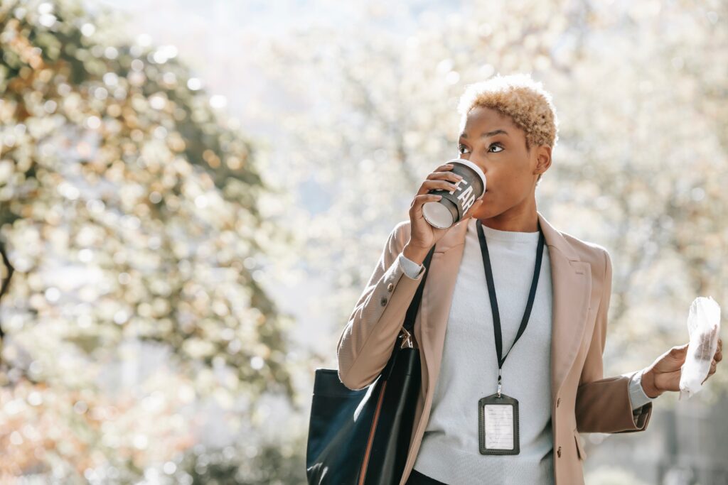 A person sips coffee from a takeout cup. In their other hand, they hold a takeout bakery item in a plastic bag. They are standing outdoors with out-of-focus trees in the background. The person is wearing a tan blazer over a white shirt, along with a badge on a lanyard. They have short cropped blonde hair and medium skin tone. Because the person appears to be taking a coffee break from work, the photo serves to illustrate a blog about employee well-being. 