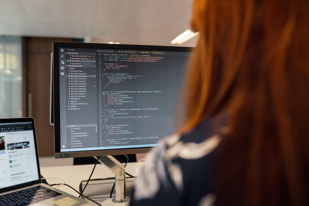 A person with long red hair faces a computer monitor and laptop displaying software data. The photo’s focal point is the data on the monitor, and its perspective is from behind the person’s left shoulder. The person is at the right of the frame, and their face is not visible. An out-of-focus office environment is visible in the background. The photo serves to illustrate a blog about how the future of personality assessment should consider the merits of traditional personality assessments along with the merits of personality assessment methods driven by AI and machine learning.